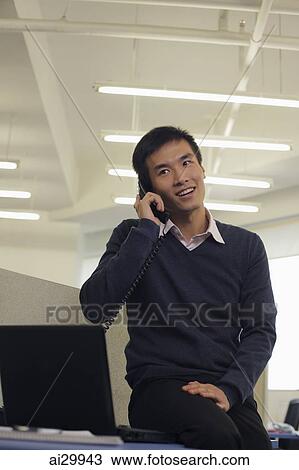 Stock Image - Young man sitting on the desk and talking on phone. Fotosearch