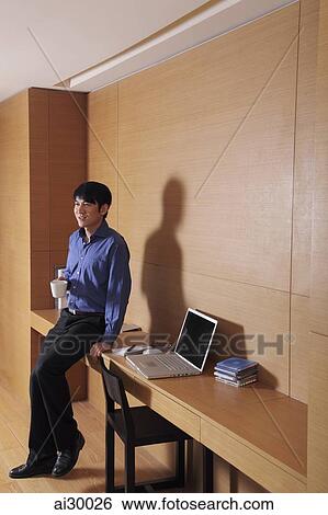 Young man sitting on the side of his desk drinking coffee View Large Photo Image Stock Photograph - Young man sitting on the side of his desk drinking coffee. Fotosearch
