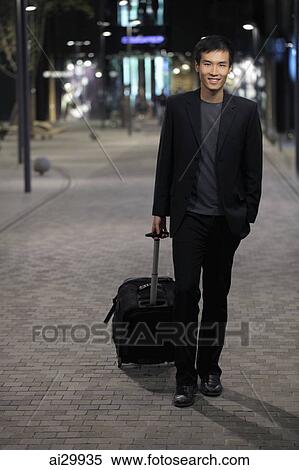 Young man walking down street pulling a suitcase View Large Photo Image Stock Photography - Young man walking down street pulling a suitcase. Fotosearch