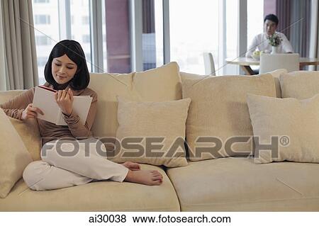 Stock Photo - Young woman reading a book on sofa with man in background. Fotosearch