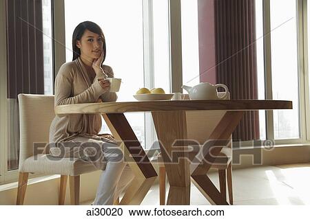 Stock Image - Young woman sitting at table drinking tea. Fotosearch