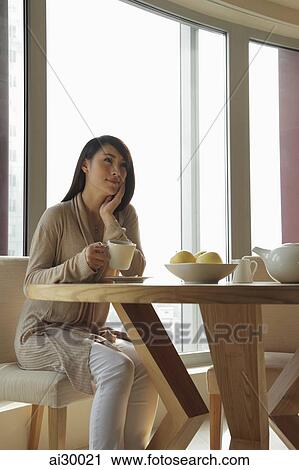 Young woman sitting at table holding a cup and looking up View Large Photo Image Stock Image - Young woman sitting at table holding a cup and looking up. Fotosearch