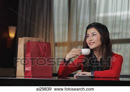 Stock Photo - Young woman sitting in restaurant with shopping bags holding cup. Fotosearch