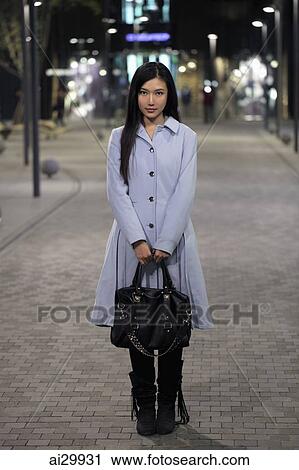 Stock Image - Young woman standing on the street holding her purse, at night. Fotosearch