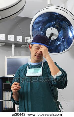 Doctor wearing scrubs standing in operating room rubbing his forehead looking tired. View Large Photo Image Stock Image - Doctor wearing scrubs standing in operating room rubbing his forehead looking tired.. Fotosearch