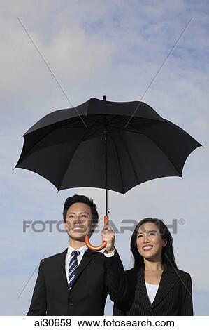 Stock Photo - Man and woman in business attire holding an umbrella and smiling. Fotosearch