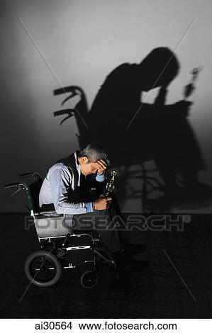 Picture - Man sitting in a wheelchair looking sad holding a trophy, his shadow on the wall. Fotosearch