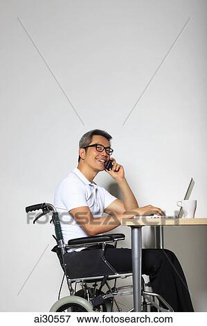 Stock Photo - Man sitting in a wheelchair using a laptop and talking on the phone. Fotosearch