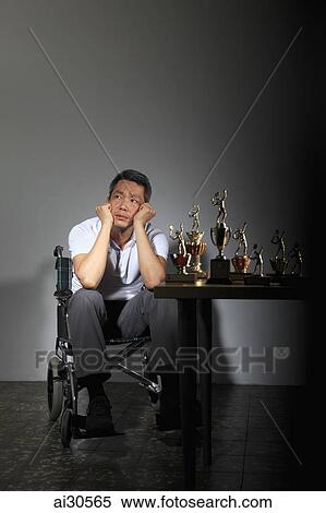 Stock Photography - Mature man sitting in a wheelchair looking sad with trophies on a table. Fotosearch