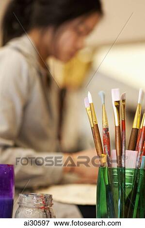 Stock Photo - Paint brushes in the foreground with young woman painting in the background. Fotosearch