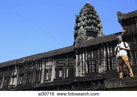 Stock Photograph - Tourist standing on the steps of Angkor Wat holding a camera, Cambodia. Fotosearch