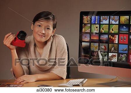 Stock Image - Woman holding a camera and sitting next to a computer of photos.. Fotosearch