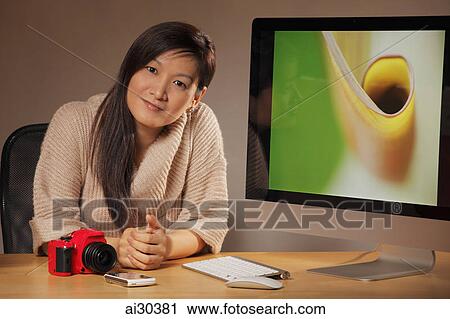 Stock Image - Woman with a camera sitting in front of a computer displaying a photograph. Fotosearch