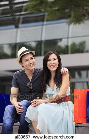 Young couple sitting outdoors with shopping bags and smiling View Large Photo Image Stock Photo - Young couple sitting outdoors with shopping bags and smiling. Fotosearch