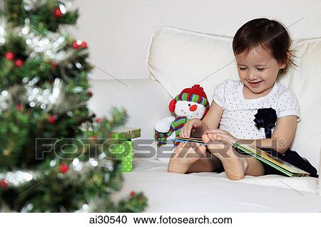 Stock Image - Young girl reading a book, Christmas tree in foreground. Fotosearch