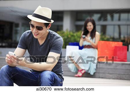 Stock Image - Young man and woman sitting outdoors looking at phones.. Fotosearch