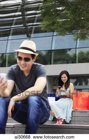 Young man and woman sitting outdoors looking at hand phones View Large Photo Image Stock Photo - Young man and woman sitting outdoors looking at hand phones. Fotosearch