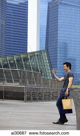 Young man holding shopping bags and looking at phone in front of buildings. View Large Photo Image Stock Photo - Young man holding shopping bags and looking at phone in front of buildings.. Fotosearch