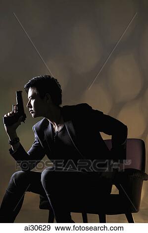 Stock Photo - Young man in profile sitting down holding a gun.. Fotosearch