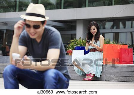 Stock Photo - Young woman and man sitting outdoors looking at phones.. Fotosearch