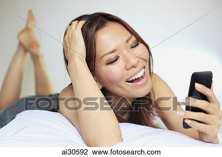 Young woman laying on a bed looking at a phone and laughing View Large Photo Image Stock Image - Young woman laying on a bed looking at a phone and laughing. Fotosearch