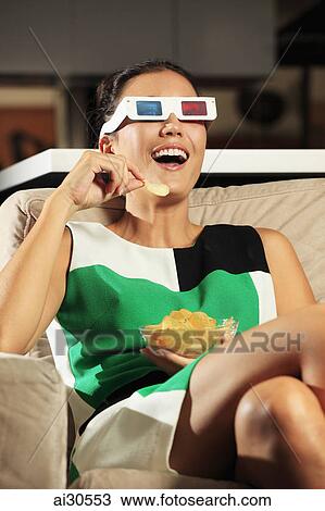 Stock Image - Young woman wearing 3D glasses and watching a movie at home and laughing.. Fotosearch