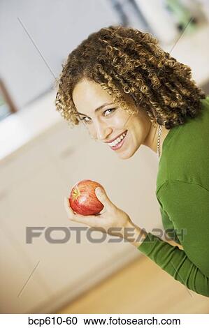 Stock Image - Portrait of woman with apple in kitchen. Fotosearch