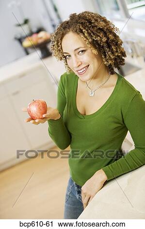 Stock Image - Portrait of woman with apple in kitchen. Fotosearch