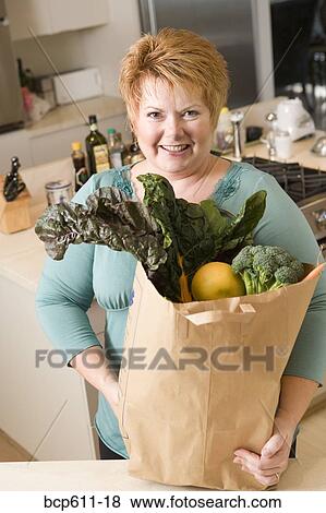 Stock Photo - Woman with bag of vegetables in kitchen. Fotosearch