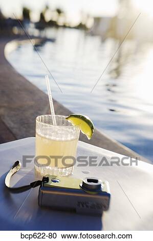 Stock Image - Margarita and camera on poolside table. Fotosearch