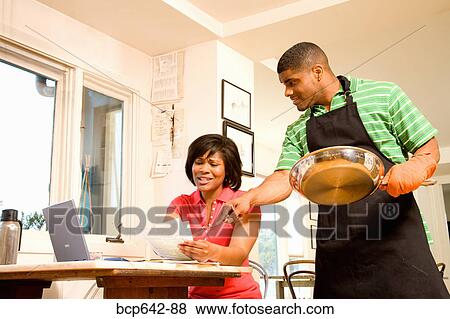Stock Photo - Man cleaning a pan as wife writes checks. Fotosearch