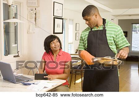 Stock Photography - Man cleaning a pan as wife writes checks. Fotosearch