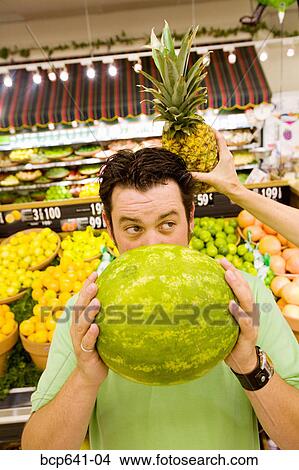 Man holding watermelon in supermarket as woman holds pineapple behind his head View Large Photo Image Picture - Man holding watermelon in supermarket as woman holds pineapple behind his head. Fotosearch
