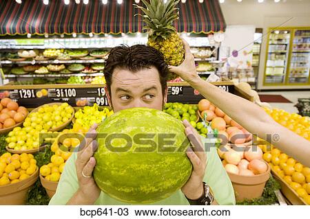 Man holding watermelon in supermarket as woman holds pineapple behind his head View Large Photo Image Stock Image - Man holding watermelon in supermarket as woman holds pineapple behind his head. Fotosearch