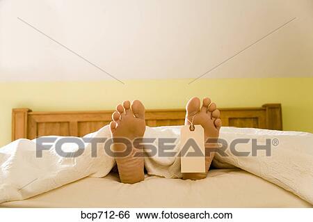 Stock Photograph - Bare feet with blank toe tag sticking out from under blanket. Fotosearch