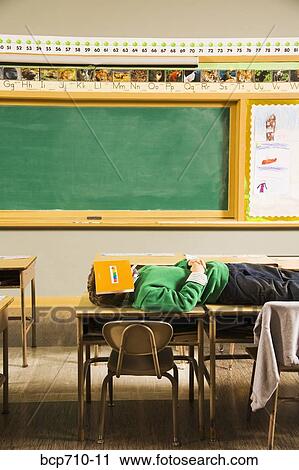 Boy laying on classroom desks with book over face View Large Photo Image Stock Image - Boy laying on classroom desks with book over face. Fotosearch