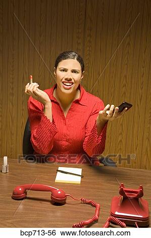 Stock Photograph - Businesswoman applying lipstick with telephone off the hook. Fotosearch