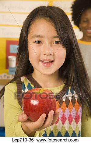 Stock Photograph - Portrait of girl holding apple in classroom. Fotosearch