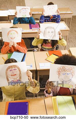 Stock Image - Students holding drawings over faces in classroom. Fotosearch