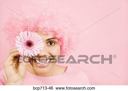 Stock Photograph - Woman wearing pink wig and holding flower over eye. Fotosearch
