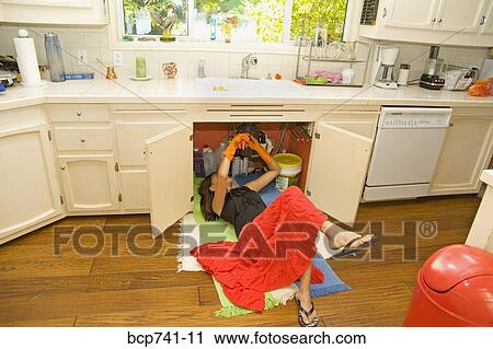 Stock Image - Woman fixing plumbing under sink. Fotosearch