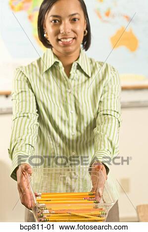 Stock Image - African female teacher holding bin of pencils. Fotosearch
