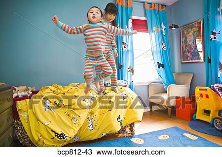 Stock Image - Asian siblings jumping on bed. Fotosearch
