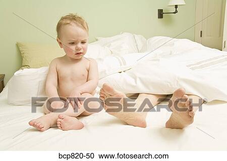 Stock Image - Curious baby looking at mother’s feet in bed. Fotosearch
