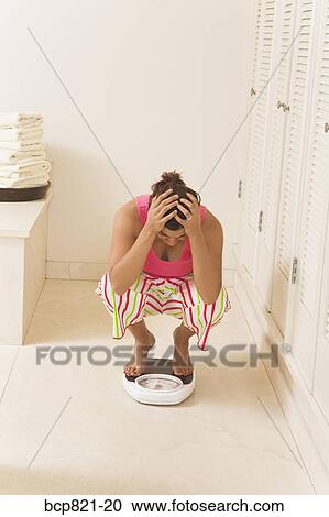 Stock Image - Frustrated woman crouching on scale. Fotosearch
