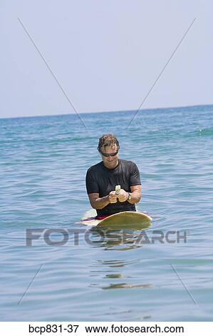 Stock Photo - Man sitting on a surfboard and text messaging. Fotosearch