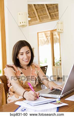 Stock Image - Woman sitting in front of a laptop and writing on a notebook. Fotosearch