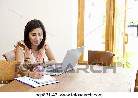 Woman sitting in front of a laptop and writing on a notebook View Large Photo Image Stock Photo - Woman sitting in front of a laptop and writing on a notebook. Fotosearch