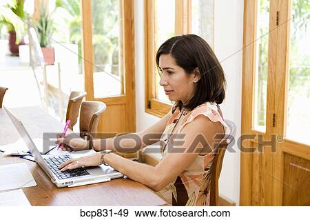 Stock Photo - Woman sitting in front of a laptop and writing on a notebook. Fotosearch