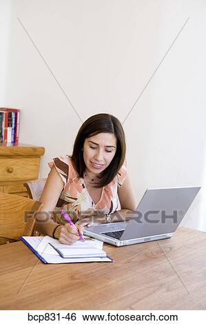Stock Photograph - Woman sitting in front of a laptop and writing on a notebook. Fotosearch